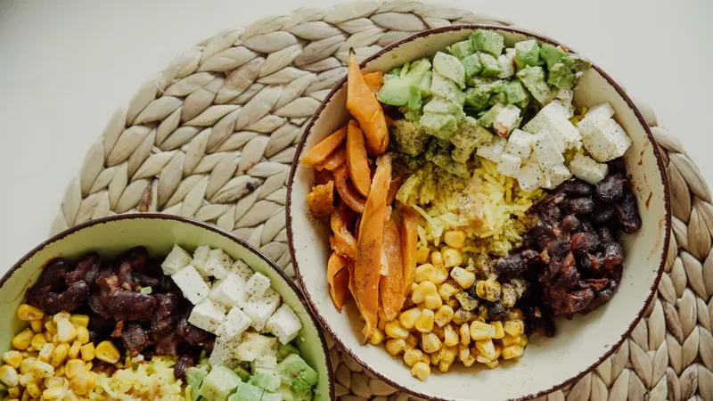 Hasır altlıkta iki kâse: avokado, tatlı patates, mısır, fasulye ve peynir; dengeli bir öğün örneği